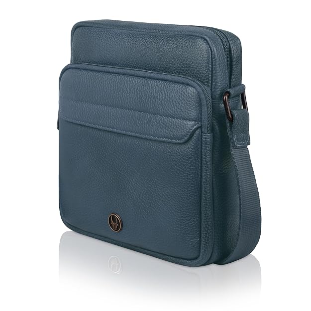 A dark teal blue pebbled leather crossbody bag with a front slip pocket, a small circular metal logo badge, and an adjustable shoulder strap, shown on a white background with a soft reflection.