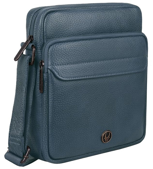 A blue pebbled leather crossbody bag with a front zippered pocket and a small circular metal logo badge, shown on a plain white background.