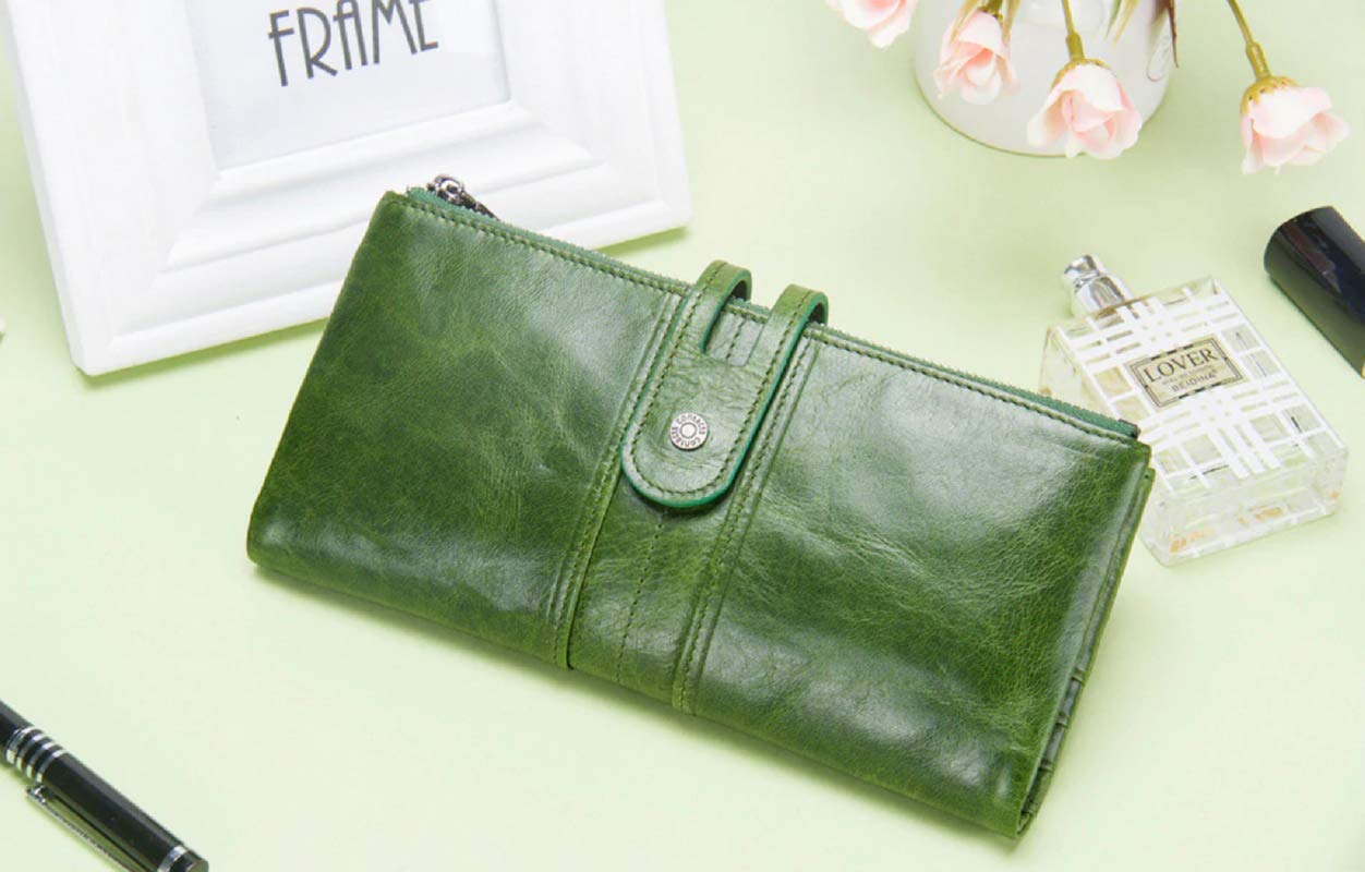 A green leather wallet with a snap-button strap closure, displayed on a pale green surface with decorative props including a pen, a perfume bottle, flowers, and a picture frame.
