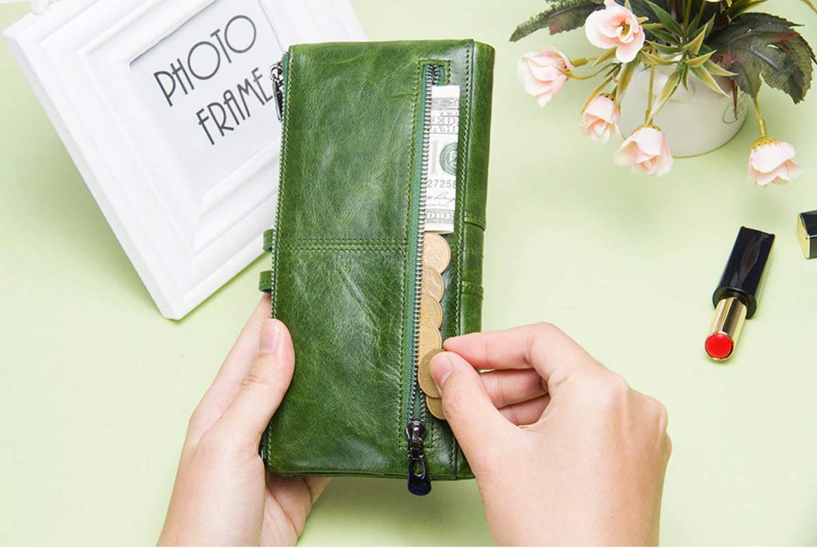 A person's hands holding an olive green leather zip-around wallet with a visible cash compartment, set against a light green background with decorative props including a white picture frame and pink flowers.