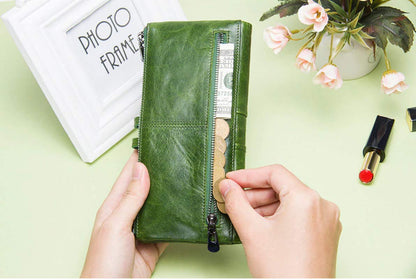 A person's hands holding an olive green leather zip-around wallet with a visible cash compartment, set against a light green background with decorative props including a white picture frame and pink flowers.